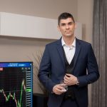 A businessman in a suit stands indoors with a stock market graph on a screen.